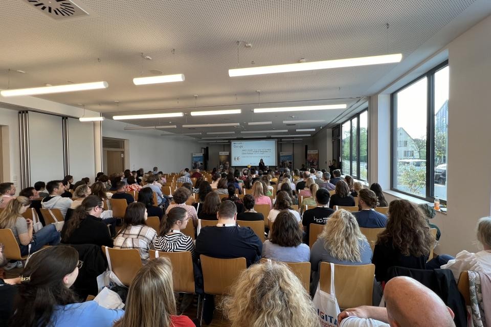 In einem Klassenzimmer sitzen die Besucherinnen und Besucher und schauen auf eine Leinwand am Ende des Raumes.