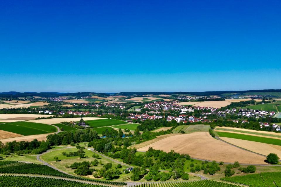 Eine Drohnenaufnahme der Gemeinde Bretzfeld. Im unteren Teil sind Weinberge mit grünen Blättern zu sehen. Danach folgen Wiesen mit Bäumen und Sträuchern. In der Mitte sind Häuser der Gemeine Bretzfeld zu sehen. Drumherum Wiesen und Äcker, teilweise grün, teilweise braun/beige. Dazwischen immer wieder Baumstreifen. In der Ferne sind Hügel zu sehen. Der Himmel ist strahlend blau.