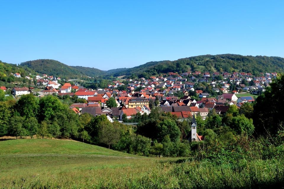 Gemeinde Mulfingen Einige Häuser zwischen grünen bewaldeten Hügeln stellt die Gemeinde Mulfingen dar. In der Mitte sticht ein gelbes älteres Bauwerk hervor. Im Vordergrund ist eine grüne Wiese und vor dem Ort ein kleiner Kapellenturm. Der Himmel ist hellblau.