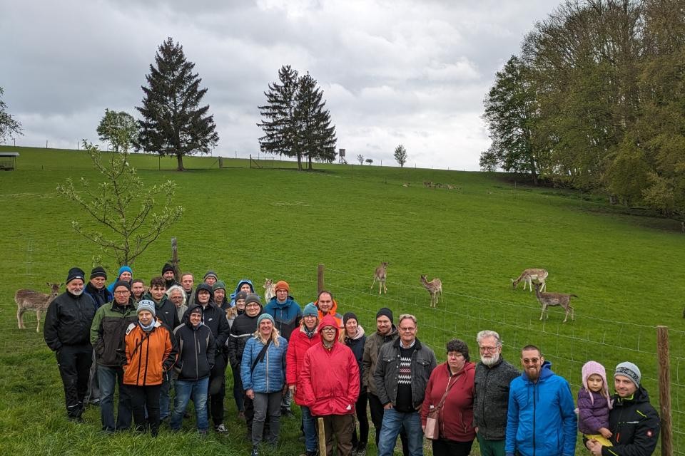 Personen stehen auf einer grünen Wiese am Hang zu einem Gruppenbild zusammen. Im Hintergrund weiden Rehe.