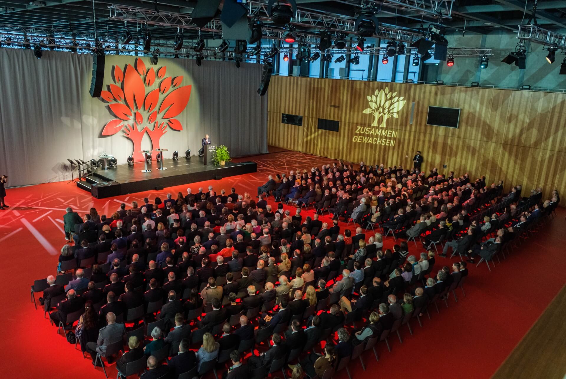 Publikum sitzt vor einer Bühne. Als Bühnenbild ist ein roter Baum der von Scheinwerfern angestrahlt wird.